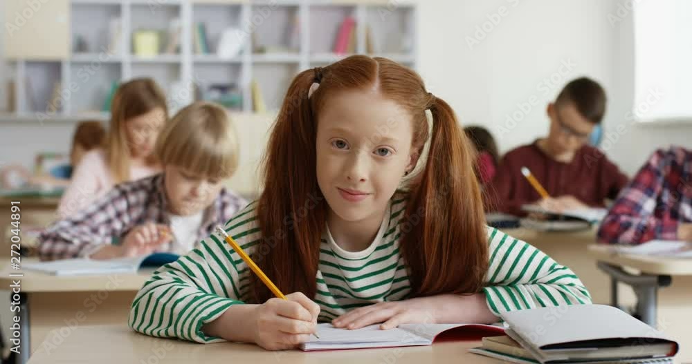 Portrait shot of the Caucasian teen red-haired schoolgirl writing in the copybook at the lesson in the classroom and smiling cheerfully to the camera. Portrait shot of the Caucasian teen red-haired schoolgirl writing in the copybook at the lesson in the classroom and smiling cheerfully to the camera.