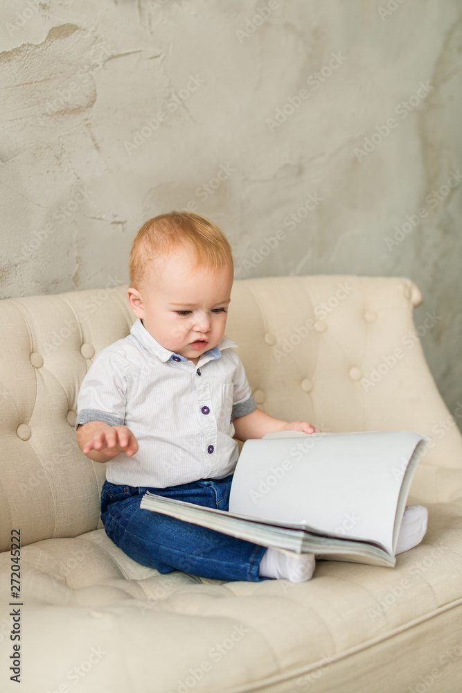 A small cute fair-haired a boy raised a looks in empty book. A red-haired boy in blue jeans and a white shirt sits on a cream sofa in the room. Vertical photography. A small cute fair-haired a boy raised a looks in empty book. A red-haired boy in blue jeans and a white shirt sits on a cream sofa in the room. Vertical photography.