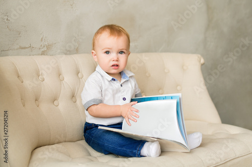 An excited little boy in blue jeans and a white shirt sits on a light sofa and holds an empty book in his hands.