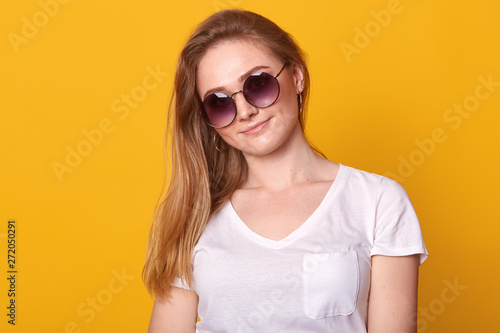 Close up portrait of cute woman with charming smile and with long blonde hairstyle  wears casual white t shirt and sunglasses  looks at camera with pleasant expression  isolated over yellow background