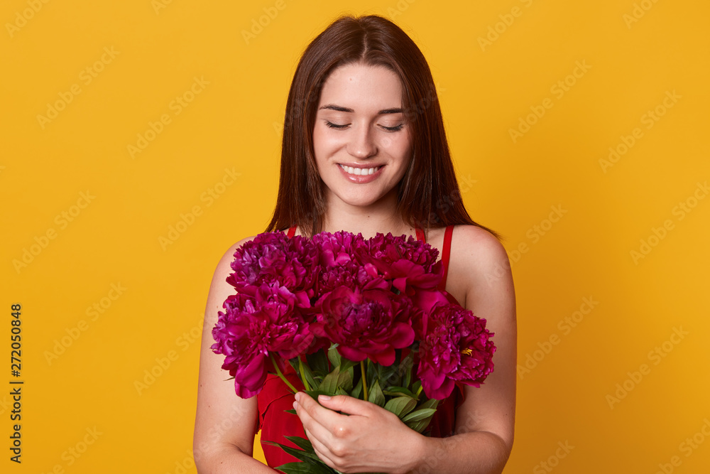 Studio shot of beautiful brunette girl holding big bouquet with burgundy peonies. Happy stylish woman with flowers, charming lady has bitrhday present, posing against yellow studio background. Studio shot of beautiful brunette girl holding big bouquet with burgundy peonies. Happy stylish woman with flowers, charming lady has bitrhday present, posing against yellow studio background.