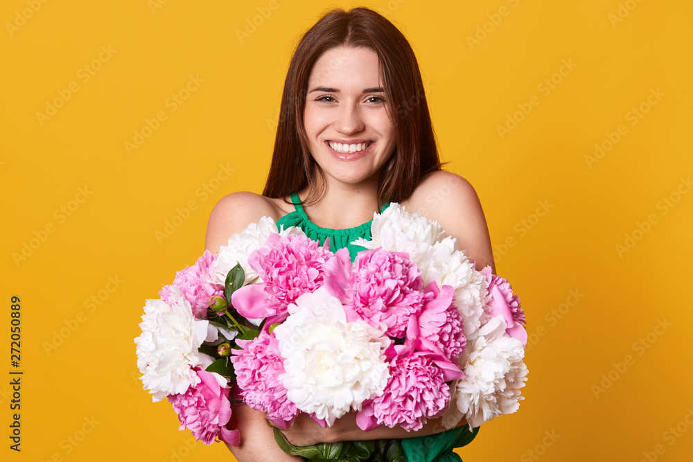 Studio shot of cheerful bright girl, looks happy, stands with toothy smile, brunette young woman holding bouquet of white and pink peonies, expresses happyness and gladness. People emotions concept. Studio shot of cheerful bright girl, looks happy, stands with toothy smile, brunette young woman holding bouquet of white and pink peonies, expresses happyness and gladness. People emotions concept.