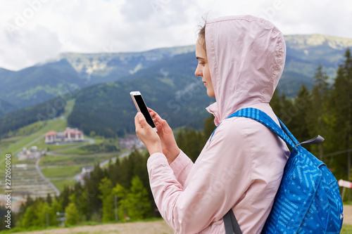 Close up profile portrait of hiker young woman with blue backpack taking photographs of beautiful landscape of mountain in spring, female wearsrose jacket with hood. Traveling and active rest concept. photo