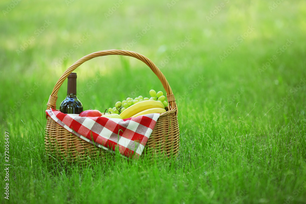Wicker basket with blanket, wine and food on green grass in park, space for text. Summer picnic Wicker basket with blanket, wine and food on green grass in park, space for text. Summer picnic