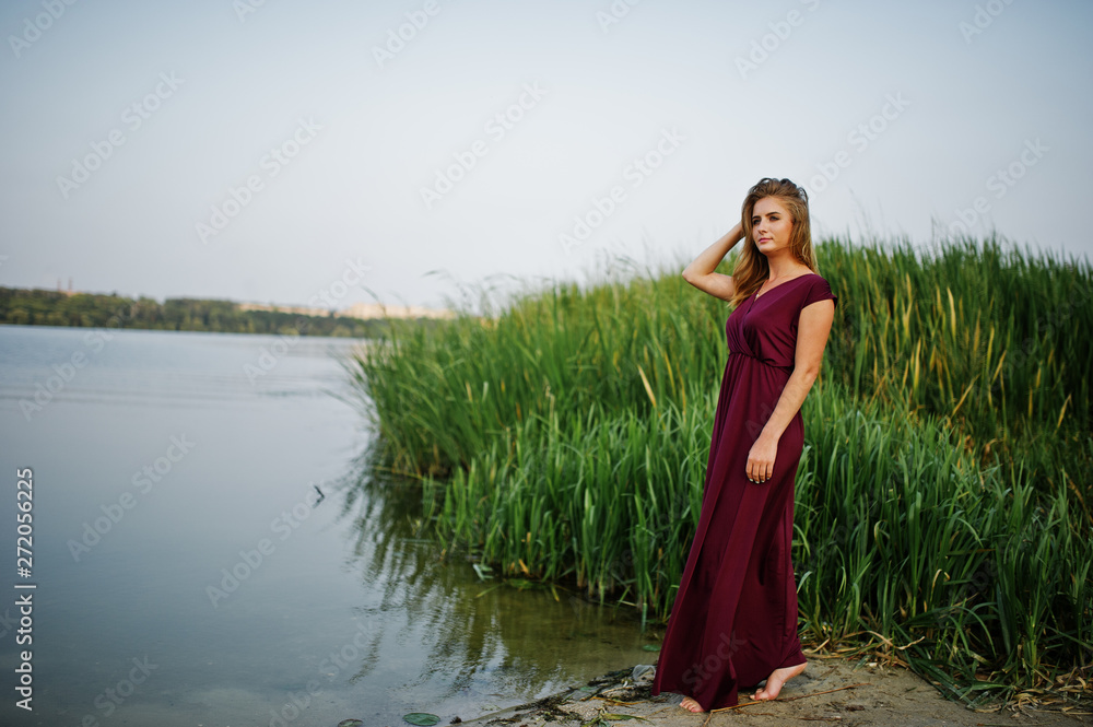 Obraz premium Blonde sensual barefoot woman in red marsala dress posing against lake with reeds.