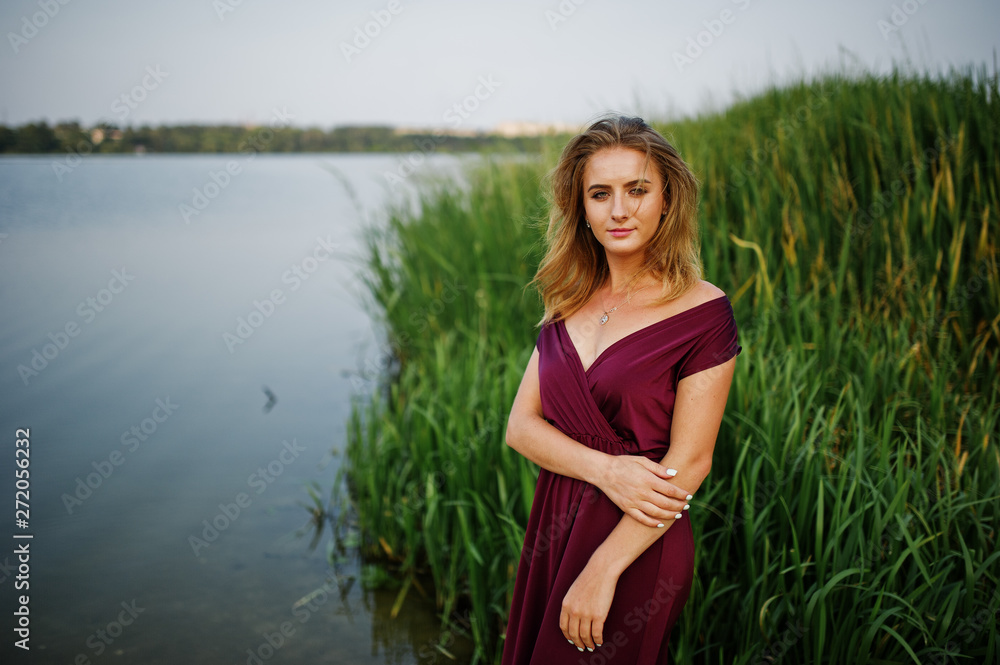 Fototapeta premium Blonde sensual woman in red marsala dress posing against lake with reeds.