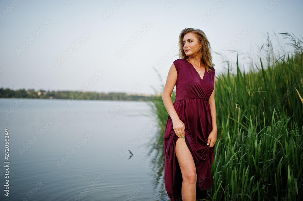 Fototapeta premium Blonde sensual woman in red marsala dress standing in water of lake with reeds.