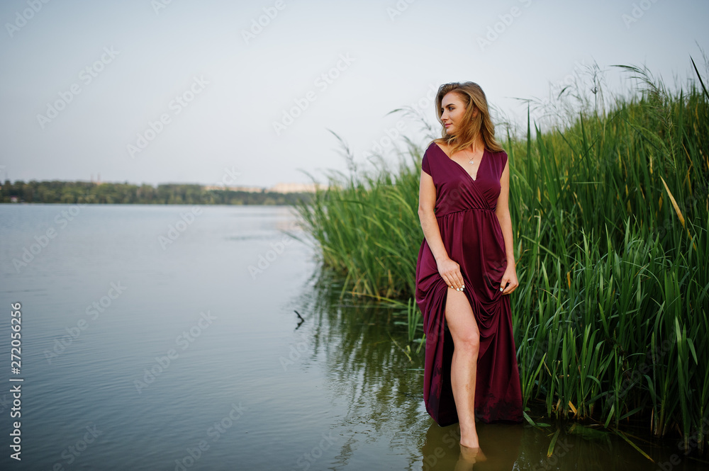 Fototapeta premium Blonde sensual woman in red marsala dress standing in water of lake with reeds.