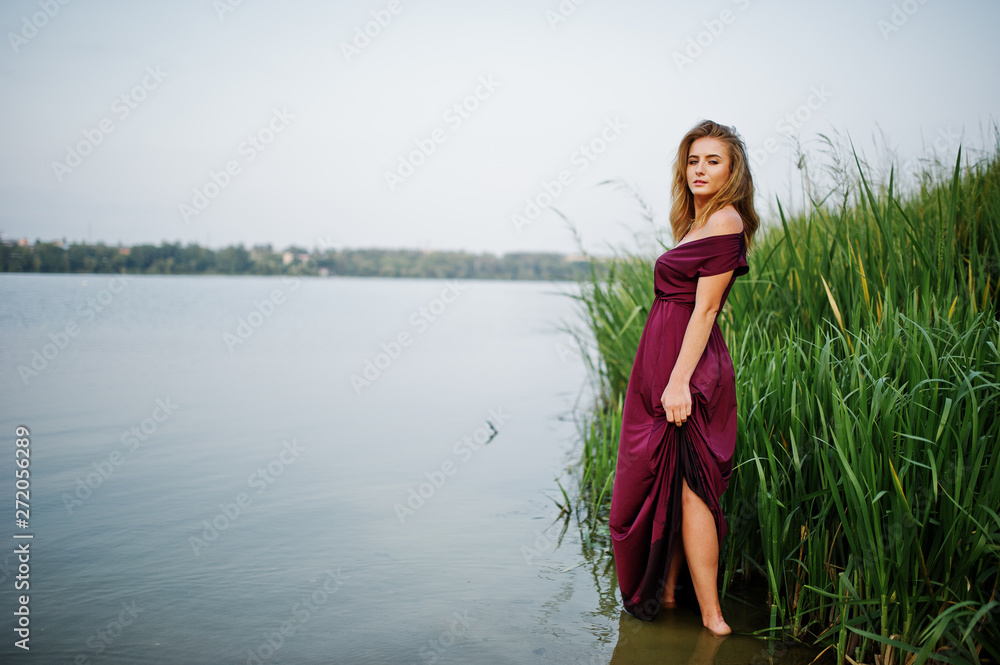 Fototapeta premium Blonde sensual woman in red marsala dress standing in water of lake with reeds.