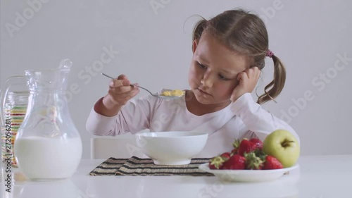 Portrait of sad small girl doesn't want to eat cornflakes with milk for breakfast. She is mixing her food with spoon sitting at table on white background.