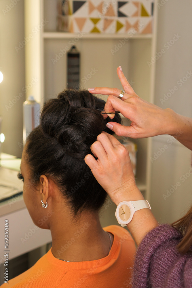 Fototapeta premium Hair stylist hands form high hairstyle on head of young African American. Pinning curls with hairpins and hair slide. Master class on creating high evening hairstyle. Training. Public lesson