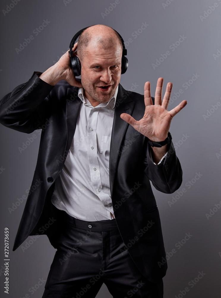 Happy emotional dancing and singing bald man listening the music in wireless headphone on dark grey background and showing five fingers on the palm. Closeup