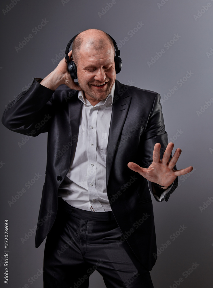 Happy emotional dancing and singing bald man listening the music in wireless headphone on dark grey background. Closeup