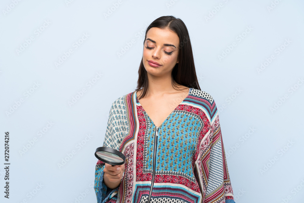 Young woman over isolated blue wall holding a magnifying glass Young woman over isolated blue wall holding a magnifying glass