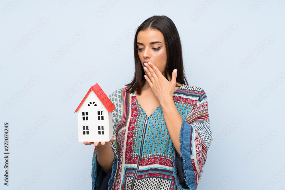 Young woman over isolated blue wall holding a little house Young woman over isolated blue wall holding a little house