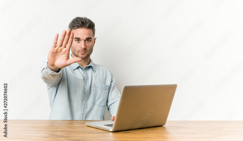 Young handsome man working with his laptop standing with outstretched hand showing stop sign, preventing you. Young handsome man working with his laptop standing with outstretched hand showing stop sign, preventing you.