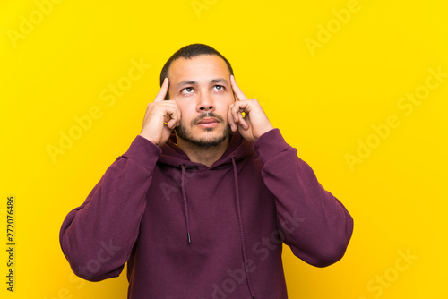 Colombian man with sweatshirt over yellow wall having doubts and thinking