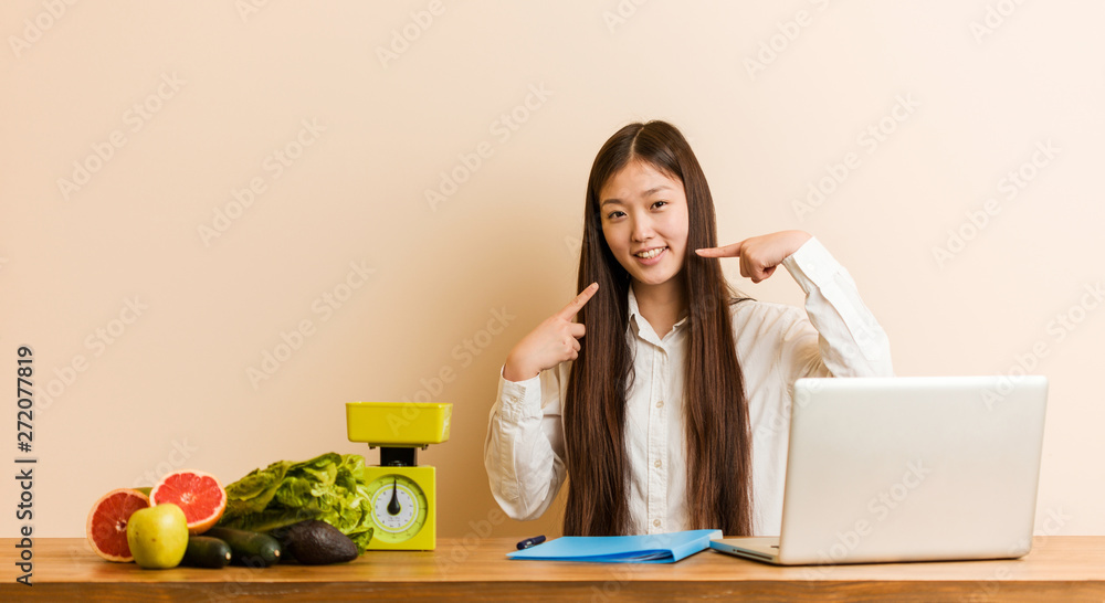Young nutritionist chinese woman working with her laptop smiles, pointing fingers at mouth. Young nutritionist chinese woman working with her laptop smiles, pointing fingers at mouth.