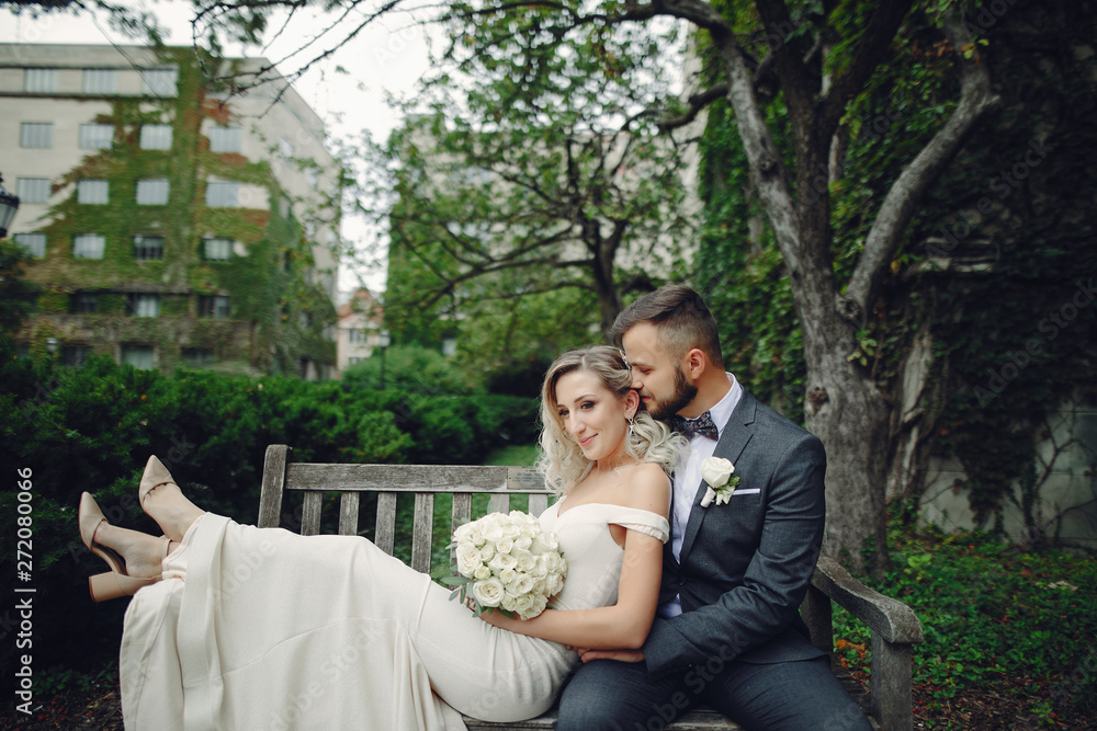 Beautiful bride in a long white dress. Handsome groom in a black suit. Couple in a summer park sitting on a bench