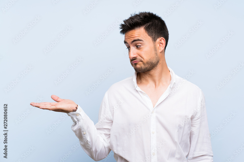 Handsome young man over isolated blue background holding copyspace with doubts Handsome young man over isolated blue background holding copyspace with doubts