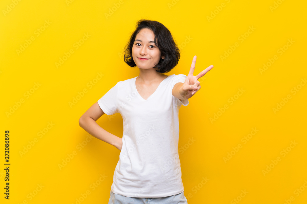 Asian young woman over isolated yellow wall smiling and showing victory sign Asian young woman over isolated yellow wall smiling and showing victory sign