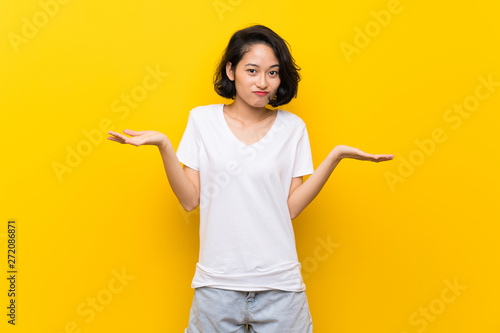 Asian young woman over isolated yellow wall having doubts while raising hands