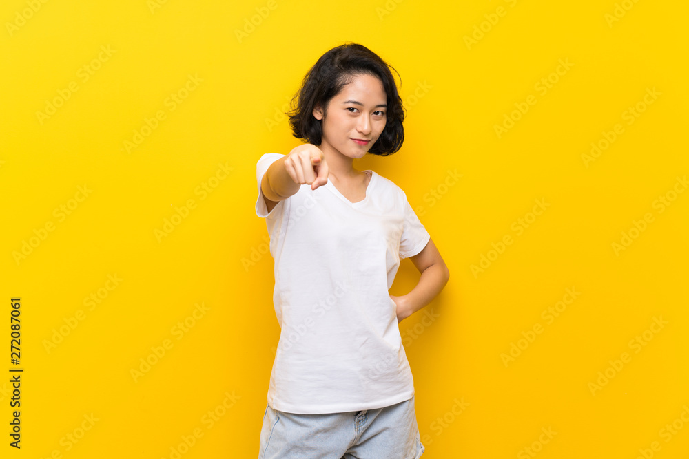 Asian young woman over isolated yellow wall points finger at you with a confident expression Asian young woman over isolated yellow wall points finger at you with a confident expression