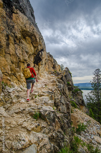 Hiker on the trail wearing red top and trainers on the trail to Inspiration point in the Grand Teton mountains NP Wyoming USA with Jenny lake below