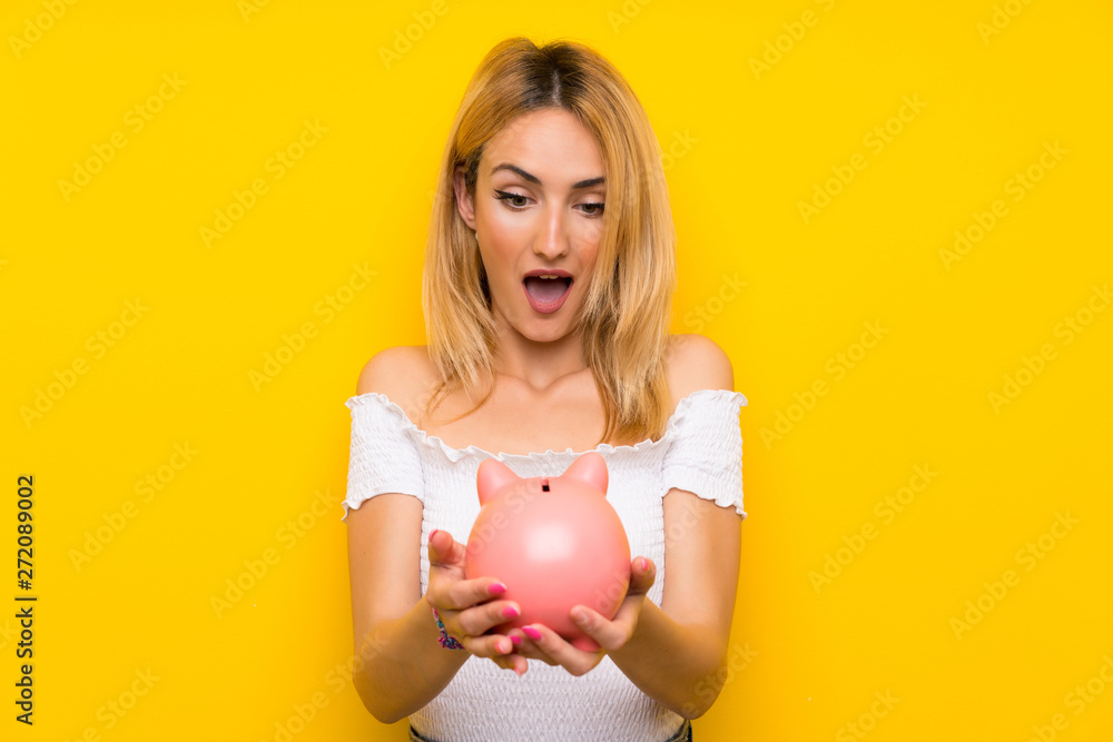 Young blonde woman over isolated yellow wall holding a big piggybank Young blonde woman over isolated yellow wall holding a big piggybank