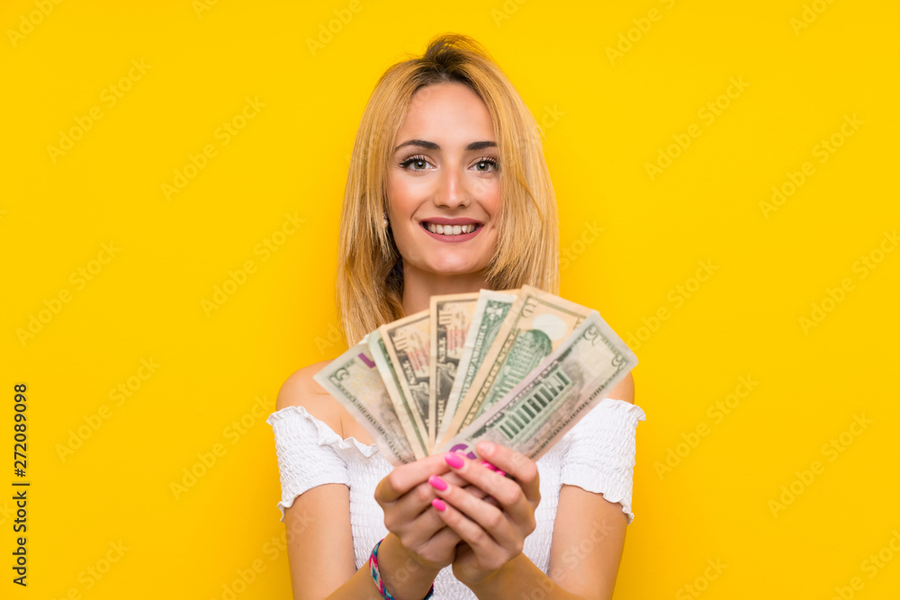 Young blonde woman over isolated yellow wall taking a lot of money