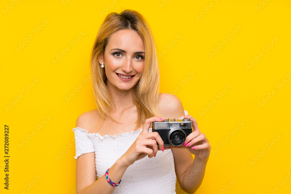 Young blonde woman over isolated yellow wall holding a camera Young blonde woman over isolated yellow wall holding a camera
