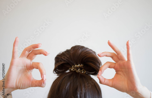 woman is doing her hair, bun for her head. Dark hair is tied with a transparent elastic band. Modern fast hairstyle. Cares about a beautiful hair. Shows ok sign