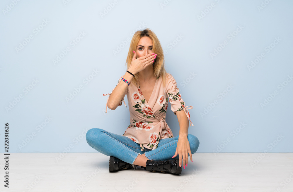Young blonde woman sitting on the floor covering mouth with hands