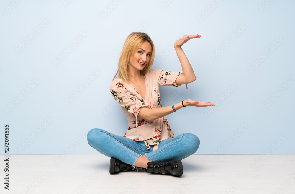Young blonde woman sitting on the floor holding copyspace to insert an ad Young blonde woman sitting on the floor holding copyspace to insert an ad