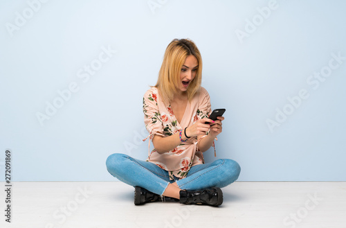 Young blonde woman sitting on the floor surprised and sending a message