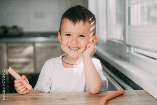 Portrait of a adorable little boy in a white t-shirt at the table eating a sausage with his hands