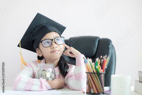 Little Asian girl wearing graduate hat hugging clear glass jar piggy bank and smile with happiness for money saving to wealthness succesful in the future of education concept select focus shallow dept photo