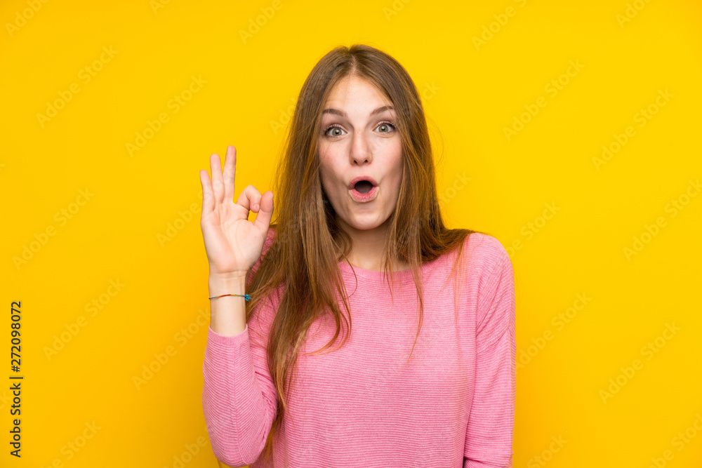 Young woman with long hair over isolated yellow wall surprised and showing ok sign Young woman with long hair over isolated yellow wall surprised and showing ok sign