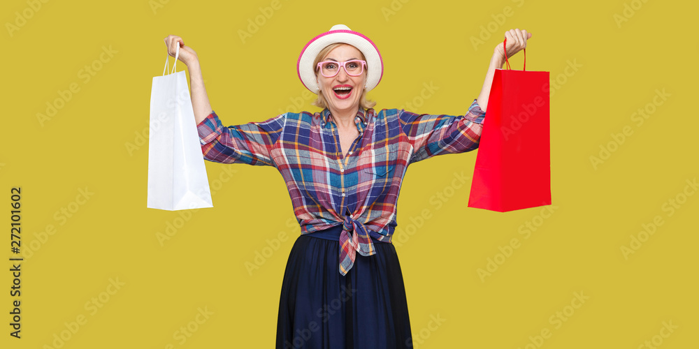 Love shopping! Satisfied modern grandma in white hat and in checkered shirt holding shopping bags and triumphing with raised arms and toothy smile. Indoor, studio shot isolated on yellow background.