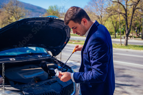 A handsome businessman wearing blue blazer lifting up the hood of his car and checking the oil level on a sunny day parked on a busy city boulevard. © qunica.com