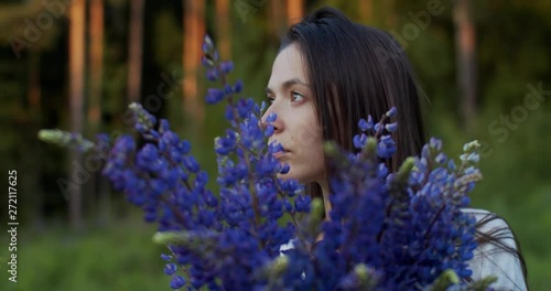 Pensive woman looks aside with bouquet of wildflowers. Side portrait of sad girl holding bunch of lupine flowers outdoors green nature shallow background slow motion. Emotions inspiration consciousnes photo