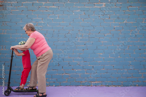 elderly woman in pink T-shirt and glasses with her little grandson is learning to ride new scooter. baby loves to spend time with her grandmother. active lifestyle concept, sport
