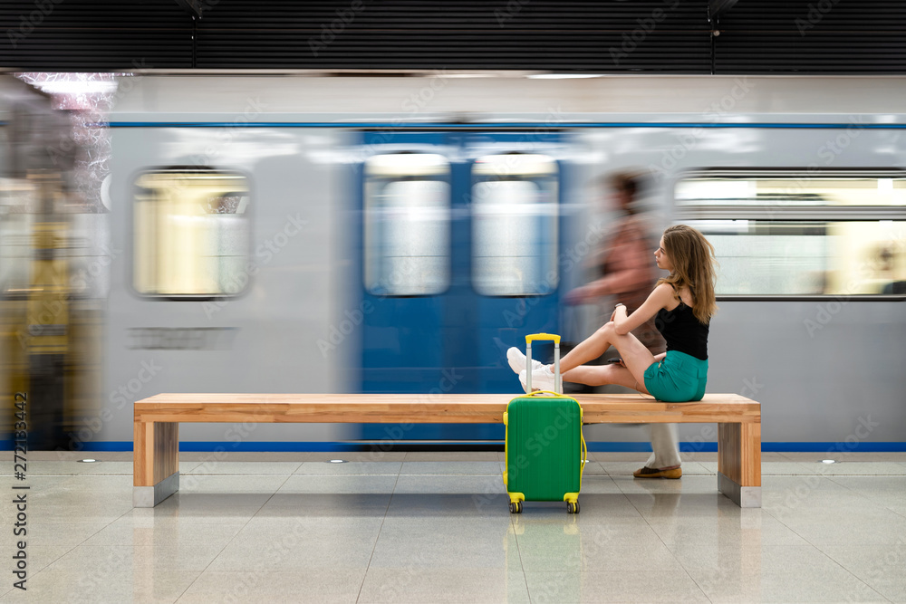 Fototapeta premium Happy girl traveler waiting for train at metro station sitting on stone bench with legs stretched out on suitcase