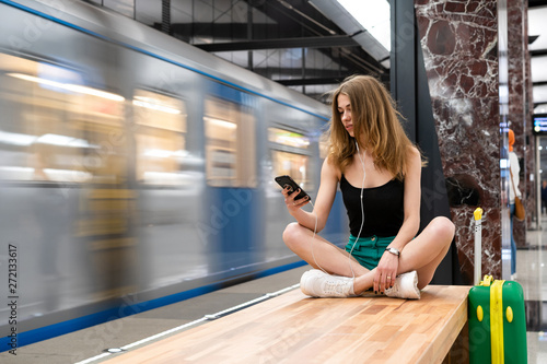 European girl sitting on a bench with a phone in her hands and in the headphones  next to a travel suitcase. It is located at the railway station or subway station. Theme of independent travel.