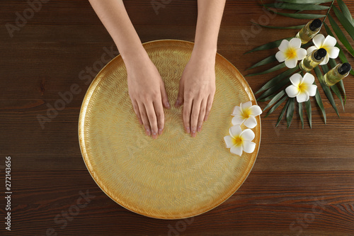 Woman soaking her hands in bowl with water and flowers on wooden table, top view. Spa treatment