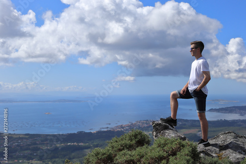 BOY WITH SUNGLASSES OBSERVING THE GALICIAN COAST FROM THE HIGH OF A MOUNTAIN ON SUMMER HOLIDAY