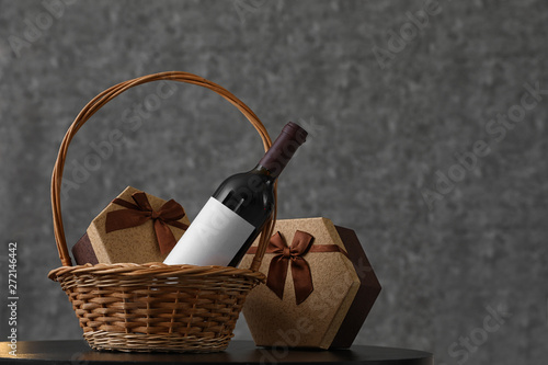 Wicker basket with bottle of wine and gift boxes on table. Space for design