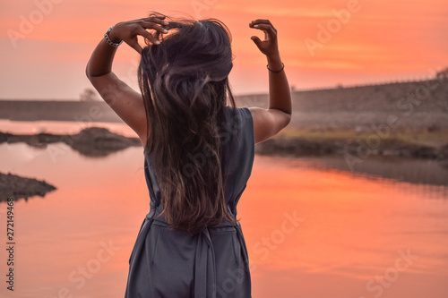 A beautiful girl with raised arms correcting hair and looking at amazing sunset at Nyari dam, Rajkot, India. 