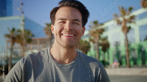 Close portrait of laughing Hispanic healthy adult standing on beach at summer day time, man joyful feeling of vacations and moment of personal happiness standing near glass edification and palms