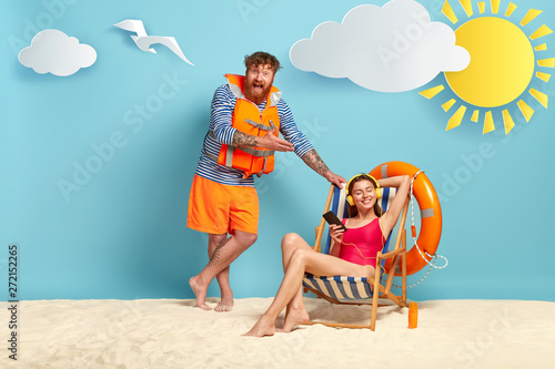 Happy man wears sailor jumper and lifejacket, stands near deckchair where relaxed woman listens music in headphones, enjoys incredible vacation, feels carefree, has lazy day. Couple have voyage photo